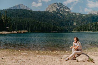 Karadağ Durmitor Milli Parkı 'ndaki göl, orman ve dağlara bakan bir kız. Turist kız gölün, kozalaklı ormanın ve dağlardaki turkuaz gölün kıyısındaki büyük taşın üzerindeki sihirli manzaranın tadını çıkarıyor. Yürüyüş.
