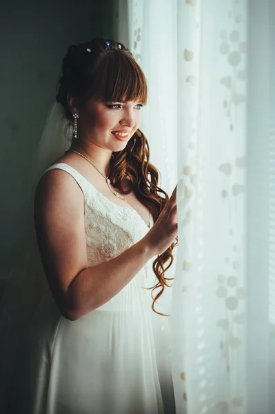 Young beautiful bride waits for groom near the window. Beautiful  young woman standing beside a large window waiting — Stock Photo, Image