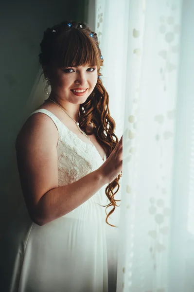 Молодая красивая невеста ждет жениха у окна. Красивая молодая женщина, стоящая у большого окна, ожидая — стоковое фото