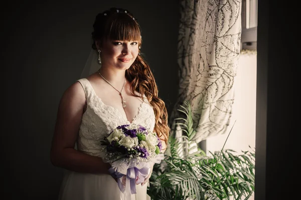 Beautiful young bride standing beside a large window waiting — Stock Photo, Image