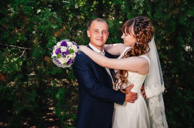 evlilik çift yeşil yaz parkta öpüşme. Gelin ve damat öpüşme, birlikte açık havada duran, yeşil ağaçlar arasında sarılma. Gelin düğün buket çiçek tutan