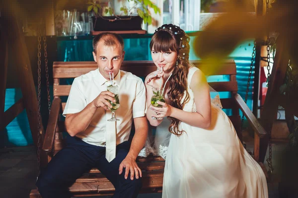 Happy bride and groom drink mojito, swinging on a swing — Stock Photo, Image