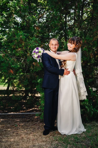 Wedding couple kissing in green summer park. bride and groom kissing, standing together outdoors, hugging among green trees. Bride holding wedding bouquet of flowers — Stock Photo, Image