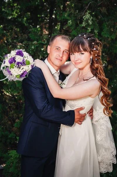 Wedding couple kissing in green summer park. bride and groom kissing, standing together outdoors, hugging among green trees. Bride holding wedding bouquet of flowers — Stock Photo, Image