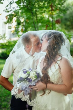 evlilik çift yeşil yaz parkta öpüşme. Gelin ve damat öpüşme, birlikte açık havada duran, yeşil ağaçlar arasında sarılma. Gelin düğün buket çiçek tutan