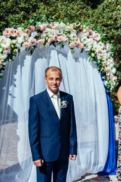 Prachtige stijlvolle rijke gelukkige bruidegom op ceremonie van het huwelijk in groene tuin in de buurt van witte boog met bloemen — Stockfoto