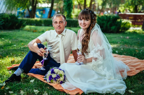 花とワインのグラスの花束と公園の緑の草の中に座ってちょうど結婚カップル — ストック写真