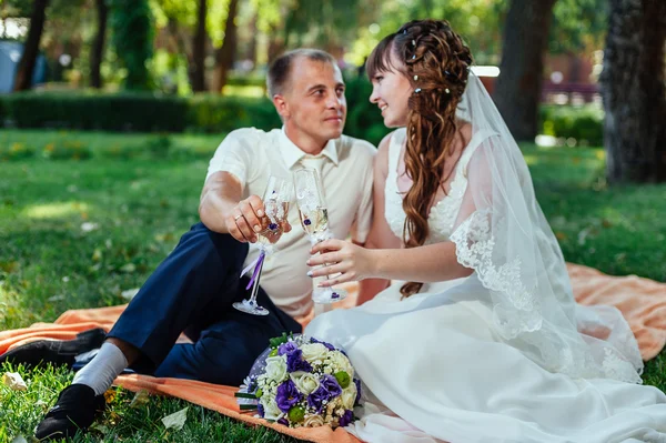 Casal acaba de se casar sentado no parque grama verde com buquê de flores e copos de vinho — Fotografia de Stock