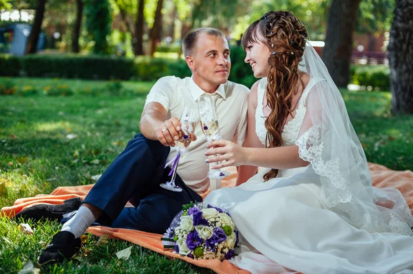 Paar, das gerade geheiratet hat, sitzt im grünen Gras des Parks mit Blumenstrauß und Weingläsern — Stockfoto
