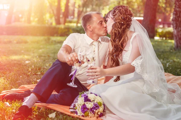 Casal acaba de se casar sentado no parque grama verde com buquê de flores e copos de vinho — Fotografia de Stock