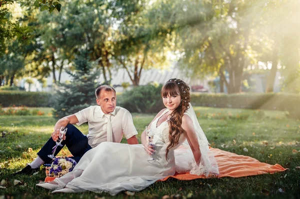 Paar, das gerade geheiratet hat, sitzt im grünen Gras des Parks mit Blumenstrauß und Weingläsern — Stockfoto