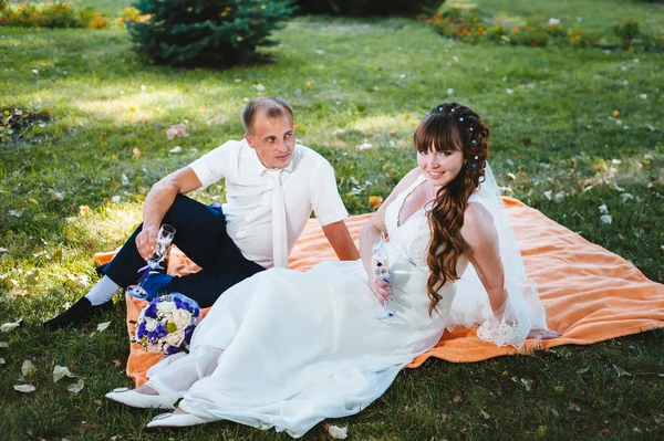 Casal acaba de se casar sentado no parque grama verde com buquê de flores e copos de vinho — Fotografia de Stock