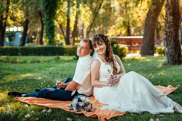 Couple just married sitting in park green grass with bouquet of flowers and wine glasses — Stock Photo, Image