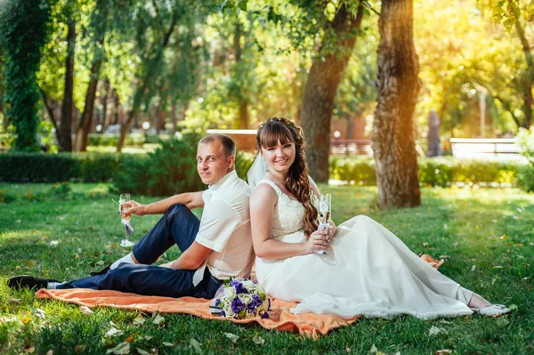 Couple just married sitting in park green grass with bouquet of flowers and wine glasses — Stock Photo, Image