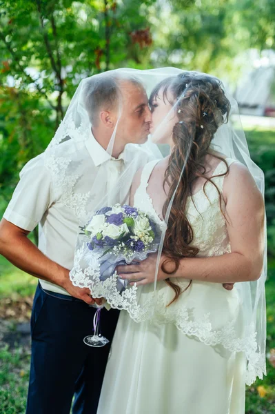 Свадебная пара целуется в зеленом летнем парке. Невеста и жених целуются, стоят вместе на улице, обнимаются между зелеными деревьями. Невеста с свадебным букетом цветов — стоковое фото