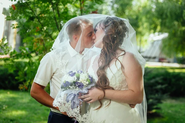 Wedding couple kissing in green summer park. bride and groom kissing, standing together outdoors, hugging among green trees. Bride holding wedding bouquet of flowers — Stock Photo, Image