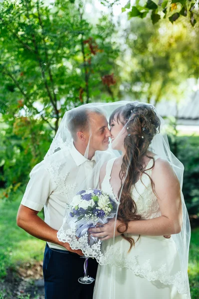 Wedding couple kissing in green summer park. bride and groom kissing, standing together outdoors, hugging among green trees. Bride holding wedding bouquet of flowers — Stock Photo, Image