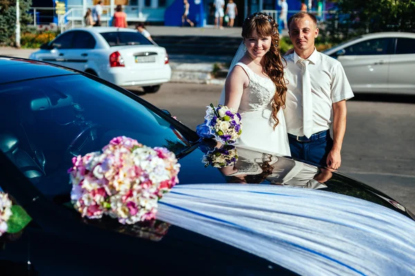 Beautiful brunette bride at white wedding with her handsome young groom in black suit stand near the car — Stock Photo, Image