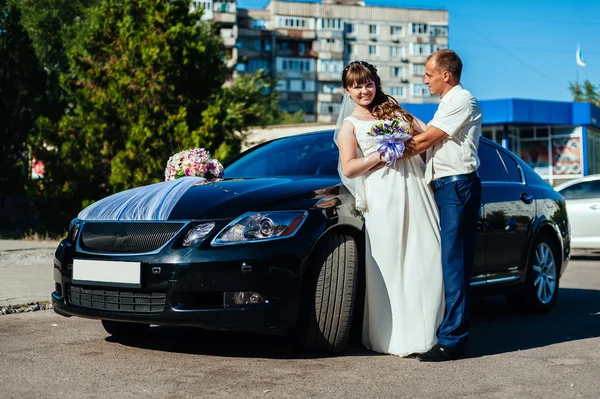 Hermosa novia morena en la boda blanca con su guapo novio joven en traje negro de pie cerca del coche — Foto de Stock