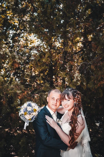 Brudparet kyssas i gröna sommaren park. Brudparet kysser, ständiga tillsammans utomhus, kramas bland gröna träd. Brud holding bröllop bukett blommor — Stockfoto