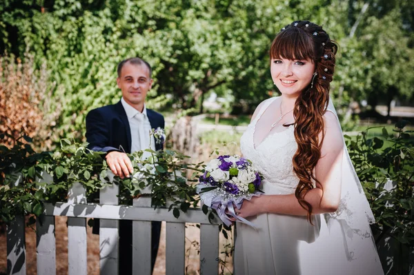 Brudparet kyssas i gröna sommaren park. Brudparet kysser, ständiga tillsammans utomhus, kramas bland gröna träd. Brud holding bröllop bukett blommor — Stockfoto