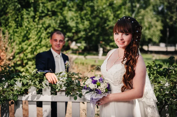 Bruidspaar zoenen in groene zomer park. bruid en bruidegom kussen, permanent samen buitenshuis, knuffelen onder de groene bomen. Bruid houden bruiloft boeket van bloemen — Stockfoto