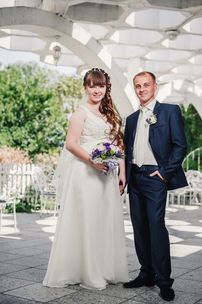 Brudparet kyssas i gröna sommaren park. Brudparet kysser, ständiga tillsammans utomhus, kramas bland gröna träd. Brud holding bröllop bukett blommor — Stockfoto