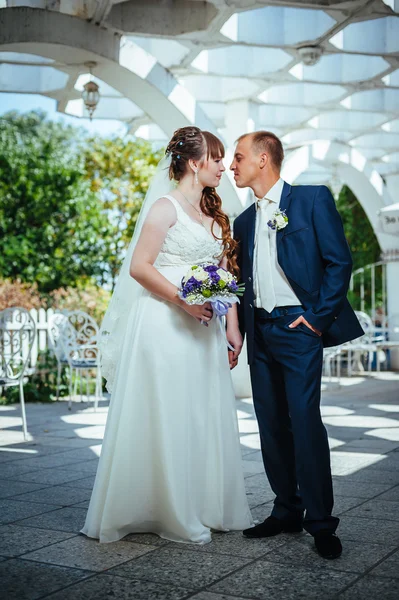 Wedding couple kissing in green summer park. bride and groom kissing, standing together outdoors, hugging among green trees. Bride holding wedding bouquet of flowers — Stock Photo, Image