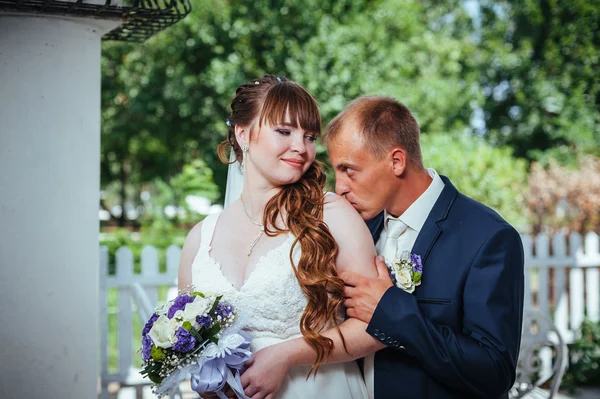Wedding couple kissing in green summer park. bride and groom kissing, standing together outdoors, hugging among green trees. Bride holding wedding bouquet of flowers — Stock Photo, Image