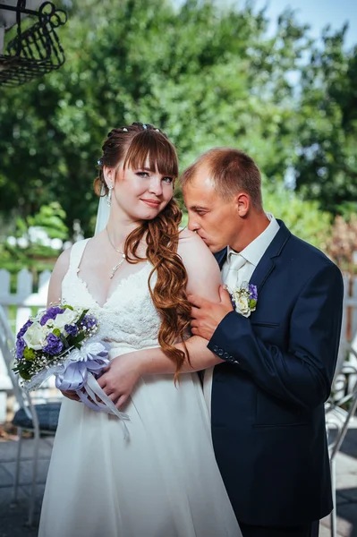 Brudparet kyssas i gröna sommaren park. Brudparet kysser, ständiga tillsammans utomhus, kramas bland gröna träd. Brud holding bröllop bukett blommor — Stockfoto