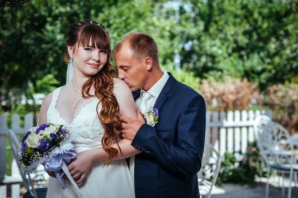 結婚式のカップルは、緑の夏の公園でキスします。花嫁と花婿のキス、一緒に屋外で立って、緑の木々 の中でハグします。花の結婚式のブーケを持って花嫁 — ストック写真