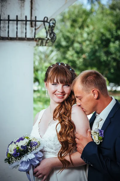 Wedding couple kissing in green summer park. bride and groom kissing, standing together outdoors, hugging among green trees. Bride holding wedding bouquet of flowers — Stock Photo, Image