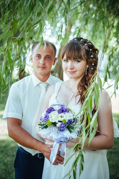 Brudparet kyssas i gröna sommaren park. Brudparet kysser, ständiga tillsammans utomhus, kramas bland gröna träd. Brud holding bröllop bukett blommor — Stockfoto