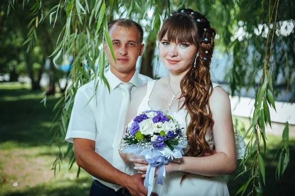 結婚式のカップルは、緑の夏の公園でキスします。花嫁と花婿のキス、一緒に屋外で立って、緑の木々 の中でハグします。花の結婚式のブーケを持って花嫁 — ストック写真