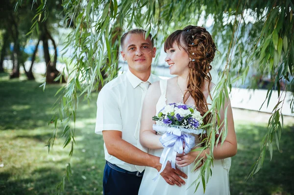 Brudparet kyssas i gröna sommaren park. Brudparet kysser, ständiga tillsammans utomhus, kramas bland gröna träd. Brud holding bröllop bukett blommor — Stockfoto