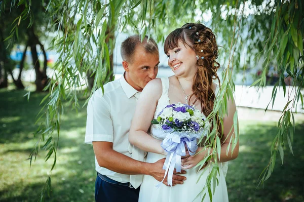 Hochzeitspaar küsst sich im grünen Sommerpark. Braut und Bräutigam küssen sich, stehen zusammen im Freien, umarmen sich unter grünen Bäumen. Braut hält Hochzeitsstrauß mit Blumen — Stockfoto