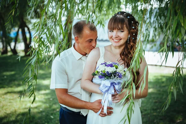 Brudparet kyssas i gröna sommaren park. Brudparet kysser, ständiga tillsammans utomhus, kramas bland gröna träd. Brud holding bröllop bukett blommor — Stockfoto