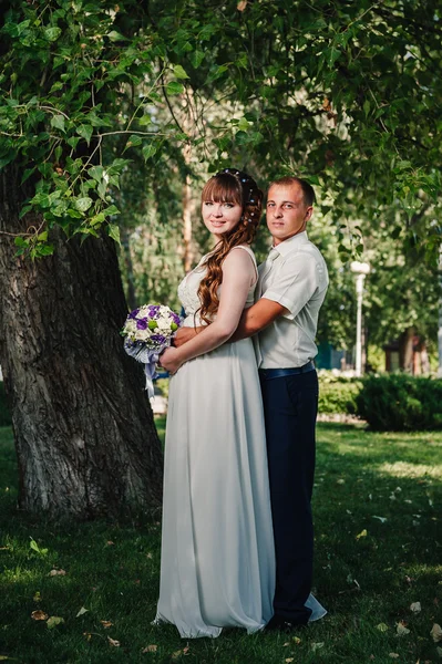 Весільна пара цілується в зеленому літньому парку. наречений і наречений цілуються, стоячи разом на відкритому повітрі, обіймаючись серед зелених дерев. Наречена тримає весільний букет квітів — стокове фото