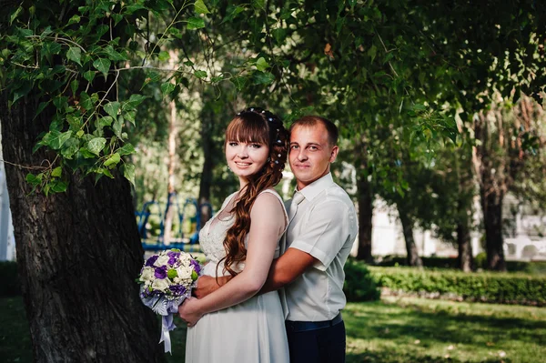Весільна пара цілується в зеленому літньому парку. наречений і наречений цілуються, стоячи разом на відкритому повітрі, обіймаючись серед зелених дерев. Наречена тримає весільний букет квітів — стокове фото