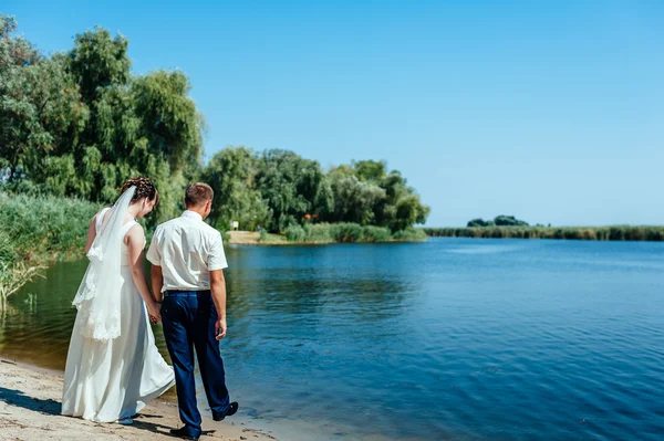 Een bruiloft door de zee. Huwelijksreis. De bruid en bruidegom knuffelen op de oever van het meer. bruid en bruidegom knuffelen op een groen meer. Bruid en bruidegom in een park. trouwjurk. Bruids bruiloft boeket van bloemen — Stockfoto