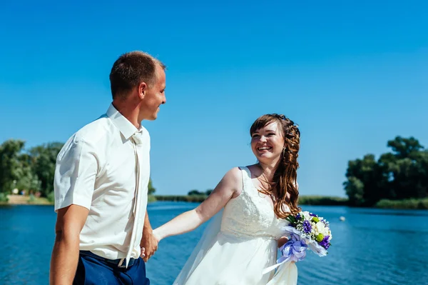 Una boda junto al mar. Luna de miel. La novia y el novio abrazándose en la orilla del lago. novio y novia abrazándose en un lago verde. Novia y novio en un parque. vestido de novia. Ramo de flores de boda nupcial —  Fotos de Stock