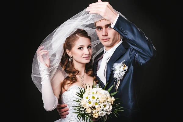 Novia y novio besándose bajo el velo sosteniendo ramo de flores en la mano. fondo negro — Foto de Stock