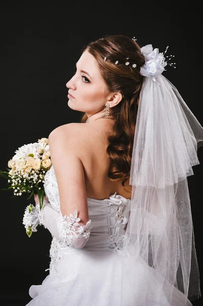 Fashion model with bridal bouquet drinking champagne and wearing wedding dress at black studio background — Stock Photo, Image