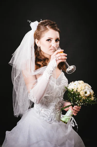 Modelo de moda com buquê de noiva bebendo champanhe e vestindo vestido de noiva no fundo do estúdio preto — Fotografia de Stock