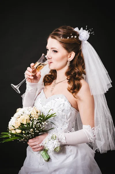 Fashion model with bridal bouquet drinking champagne and wearing wedding dress at black studio background — Stock Photo, Image