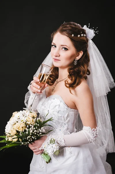 Fashion model with bridal bouquet drinking champagne and wearing wedding dress at black studio background — Stock Photo, Image