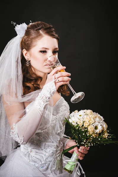 Fashion model with bridal bouquet drinking champagne and wearing wedding dress at black studio background Stock Photo