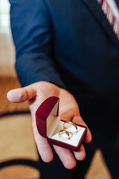 Groom holds a jewelry gift box with gold wedding rings. Man in suit and tie holding engagement ring