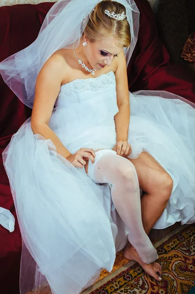 Strumpfband am Bein der Braut, schlanke sexy Braut im Brautluxuskleid, die ihren seidenen Strumpfband mit goldener Schleife zeigt. Frauen haben letzte Vorbereitungen für die Trauung. Hochzeitstag — Stockfoto