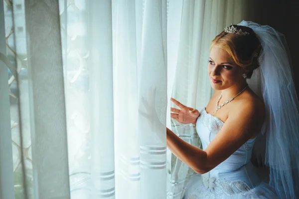 Beautiful young bride standing beside a large window waiting — Stock Photo, Image